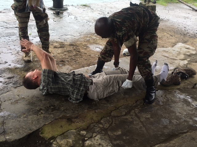 A military training or medical exercise scenario is depicted outdoors on a wet concrete surface. A soldier in camouflage uniform and black gloves is attending to a civilian-clothed person lying on the ground, likely simulating casualty care or a search procedure. At least one additional uniformed individual is visible in the background.