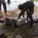 A military training or medical exercise scenario is depicted outdoors on a wet concrete surface. A soldier in camouflage uniform and black gloves is attending to a civilian-clothed person lying on the ground, likely simulating casualty care or a search procedure. At least one additional uniformed individual is visible in the background.