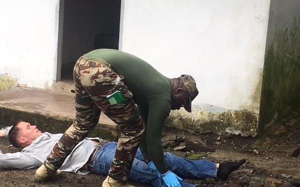 A person in military camouflage uniform with a green flag patch is bending over and attending to a person lying on the ground outdoors. The standing individual is wearing blue latex gloves, suggesting a medical or first-aid context. The scene appears to be a training exercise or emergency response scenario near a white building.