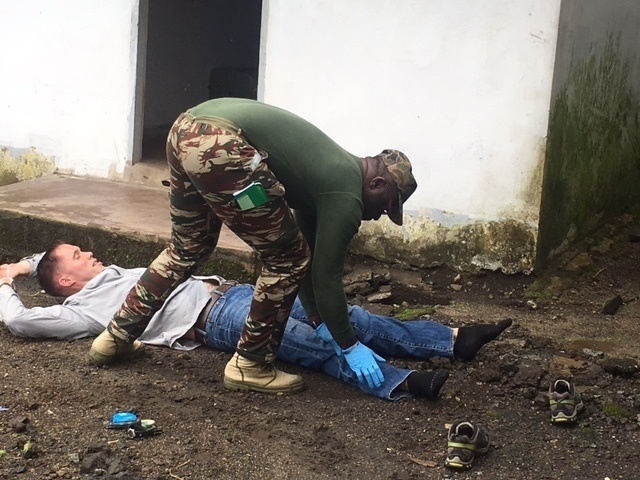 A person in military camouflage uniform with a green flag patch is bending over and attending to a person lying on the ground outdoors. The standing individual is wearing blue latex gloves, suggesting a medical or first-aid context. The scene appears to be a training exercise or emergency response scenario near a white building.