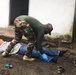 A person in military camouflage uniform with a green flag patch is bending over and attending to a person lying on the ground outdoors. The standing individual is wearing blue latex gloves, suggesting a medical or first-aid context. The scene appears to be a training exercise or emergency response scenario near a white building.
