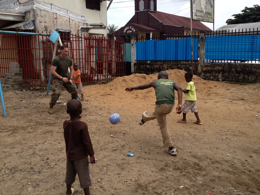 A U.S. Marine in military uniform is playing soccer with several local children in what appears to be an urban area in West Africa, likely Liberia based on contextual clues. The game is taking place on a dirt surface near a red metal fence, with a church building visible in the background. The scene depicts a community engagement or humanitarian interaction between military personnel and local youth.