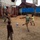 A U.S. Marine in military uniform is playing soccer with several local children in what appears to be an urban area in West Africa, likely Liberia based on contextual clues. The game is taking place on a dirt surface near a red metal fence, with a church building visible in the background. The scene depicts a community engagement or humanitarian interaction between military personnel and local youth.