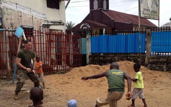 A U.S. Marine in military uniform is playing soccer with several local children in what appears to be an urban area in West Africa, likely Liberia based on contextual clues. The game is taking place on a dirt surface near a red metal fence, with a church building visible in the background. The scene depicts a community engagement or humanitarian interaction between military personnel and local youth.