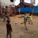 A U.S. Marine in military uniform is playing soccer with several local children in what appears to be an urban area in West Africa, likely Liberia based on contextual clues. The game is taking place on a dirt surface near a red metal fence, with a church building visible in the background. The scene depicts a community engagement or humanitarian interaction between military personnel and local youth.