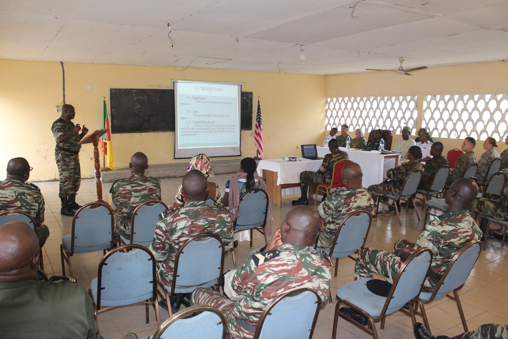 A military briefing or training session is taking place in a classroom-style setting. A uniformed officer stands at a podium presenting to an audience of military personnel seated in rows of chairs, with a projected presentation screen visible. Both African military personnel in camouflage uniforms and what appear to be Western military advisors are present, along with an American flag and what appears to be a Senegalese flag.
