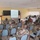 A military briefing or training session is taking place in a classroom-style setting. A uniformed officer stands at a podium presenting to an audience of military personnel seated in rows of chairs, with a projected presentation screen visible. Both African military personnel in camouflage uniforms and what appear to be Western military advisors are present, along with an American flag and what appears to be a Senegalese flag.