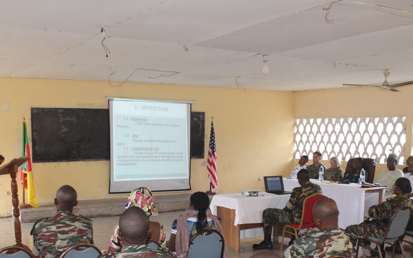 A military briefing or training session is taking place in a classroom-style setting. A uniformed officer stands at a podium presenting to an audience of military personnel seated in rows of chairs, with a projected presentation screen visible. Both African military personnel in camouflage uniforms and what appear to be Western military advisors are present, along with an American flag and what appears to be a Senegalese flag.