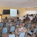 A military briefing or training session is taking place in a classroom-style setting. A uniformed officer stands at a podium presenting to an audience of military personnel seated in rows of chairs, with a projected presentation screen visible. Both African military personnel in camouflage uniforms and what appear to be Western military advisors are present, along with an American flag and what appears to be a Senegalese flag.