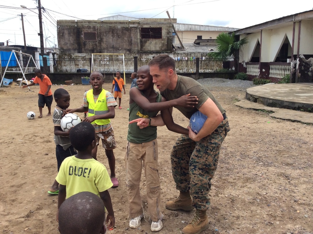A person in U.S. Marine Corps camouflage uniform interacts with a group of children on a dirt outdoor court or field. The marine is bending down and engaging closely with one child while others gather around, with a soccer ball visible nearby. The setting appears to be an urban area in a developing region, suggesting a community outreach or humanitarian engagement activity.