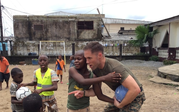 A person in U.S. Marine Corps camouflage uniform interacts with a group of children on a dirt outdoor court or field. The marine is bending down and engaging closely with one child while others gather around, with a soccer ball visible nearby. The setting appears to be an urban area in a developing region, suggesting a community outreach or humanitarian engagement activity.