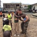 A person in U.S. Marine Corps camouflage uniform interacts with a group of children on a dirt outdoor court or field. The marine is bending down and engaging closely with one child while others gather around, with a soccer ball visible nearby. The setting appears to be an urban area in a developing region, suggesting a community outreach or humanitarian engagement activity.