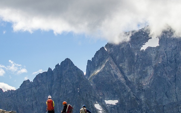 Three hikers stand on a rocky alpine ridge gazing toward a dramatic range of jagged, snow-patched mountain peaks partially shrouded in clouds. The foreground is dominated by a boulder-strewn slope with sparse low vegetation, while the background features steep granite cliffs and residual snowfields. The image captures a clear high-altitude wilderness setting under a partly cloudy sky.