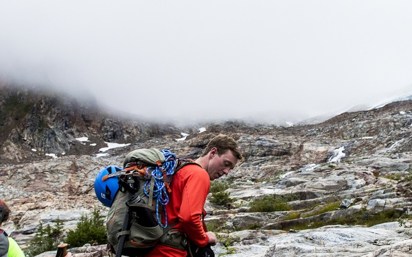 Three hikers are traversing a rocky alpine terrain in overcast, foggy conditions. The group appears to be on a technical mountaineering or glacier approach route, carrying heavy packs loaded with climbing gear. Snow patches and a possible waterfall are visible in the background, suggesting high elevation.