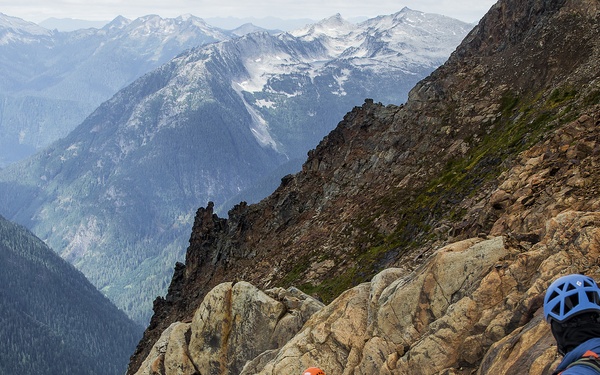Three mountaineers are traversing a rocky, snow-covered alpine ridge at high elevation, connected by a red rope for safety. They are equipped with climbing gear including helmets, backpacks, and ice axes, moving along a technical mountain route. A dramatic valley with forested slopes and snow-capped peaks stretches into the background.