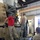 The image shows military personnel and a civilian technician working on a large multi-barrel rotary cannon system (consistent with a Phalanx CIWS or M168 Vulcan-type weapon) mounted on a trailer inside an indoor maintenance facility. Two individuals are standing on the trailer platform directly interacting with the weapon system, while a third person observes from ground level. The setting appears to be a military maintenance bay or armory.