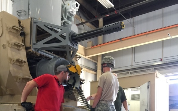 The image shows military personnel and a civilian technician working on a large multi-barrel rotary cannon system (consistent with a Phalanx CIWS or M168 Vulcan-type weapon) mounted on a trailer inside an indoor maintenance facility. Two individuals are standing on the trailer platform directly interacting with the weapon system, while a third person observes from ground level. The setting appears to be a military maintenance bay or armory.