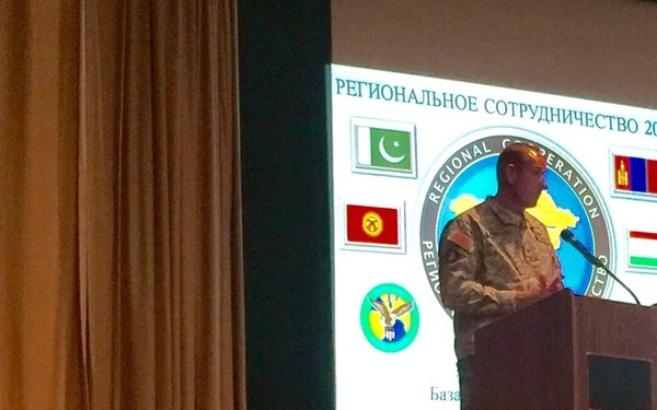 Two U.S. Army soldiers are participating in a briefing or presentation for 'Regional Cooperation 2016,' a multinational military exercise held at Camp Edwards, Massachusetts, from 16-28 September 2016. One soldier stands at a wooden podium addressing an audience, while another stands to the left holding papers. The presentation slide is displayed in both English and Russian (Cyrillic), featuring flags of participating nations including Pakistan, Mongolia, Kyrgyzstan, Tajikistan, and the United States.