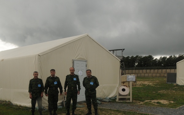 A group of five individuals, four wearing military camouflage uniforms with blue badges and one in civilian clothing, are gathered in front of a large white canvas tent structure at what appears to be a military or training compound. The facility includes gravel pathways, additional tent structures, a perimeter wall with razor wire, and a watchtower visible in the background. The sky is overcast with dark storm clouds.