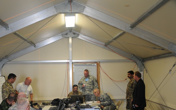 A group of approximately 8-9 individuals, most wearing military uniforms of varying camouflage patterns, are gathered inside a temporary military command tent or field operations center. They are clustered around folding tables equipped with laptops, communications equipment, and phones, suggesting a joint operations or planning session. The setting appears to be a multinational military exercise or coordination meeting.