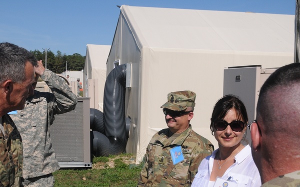 A group of U.S. Army personnel and one civilian woman are gathered outdoors near military field tents or temporary structures. Three soldiers are in camouflage uniforms, with one appearing to salute or gesture, while a civilian woman in a white blouse and sunglasses stands among them. The setting appears to be a military installation or training area with modular tent structures and HVAC equipment visible in the background.
