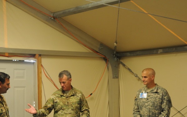 Four military personnel in different camouflage uniforms are gathered around a table equipped with laptops and communications equipment inside a temporary military command tent structure. One individual in the center appears to be briefing or explaining something to the others, gesturing with his hands. The setting suggests a multinational military exercise or coordination meeting.