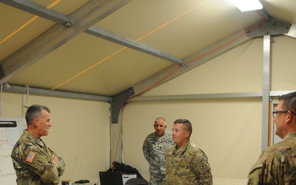 Four U.S. Army soldiers in MultiCam/OCP camouflage uniforms are gathered inside a military tent or temporary structure, appearing to be in a briefing or planning session. A whiteboard with handwritten notes is visible to the left, and folding tables with laptops and equipment are set up in the background. The setting suggests a forward operating base or tactical operations center environment.