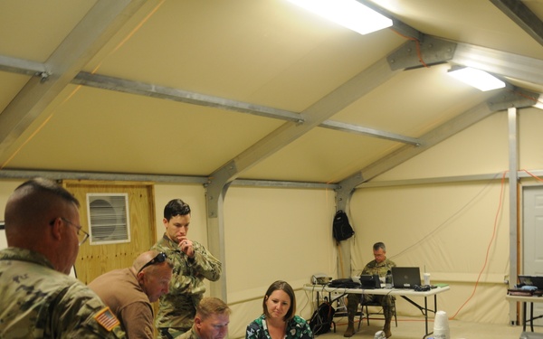 A group of military personnel and at least one civilian are gathered inside a large military-style tent or expeditionary shelter, working around tables equipped with laptops and other equipment. The scene appears to be a command post or operations center during a training exercise or real-world mission. Personnel from multiple branches are visible, including Army soldiers in ACU/OCP uniforms and a Navy sailor in NWU (Navy Working Uniform).