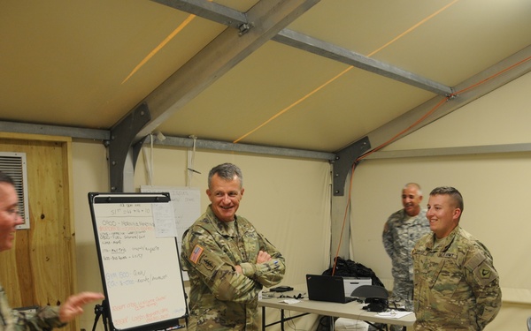 A group of U.S. Army soldiers in MultiCam/OCP camouflage uniforms are gathered in what appears to be a forward operating base or temporary command post tent structure. The soldiers appear to be engaged in an informal briefing or discussion, with one senior soldier standing with arms crossed and smiling. The setting includes a whiteboard with notes, laptops, and office supplies consistent with a tactical operations center.