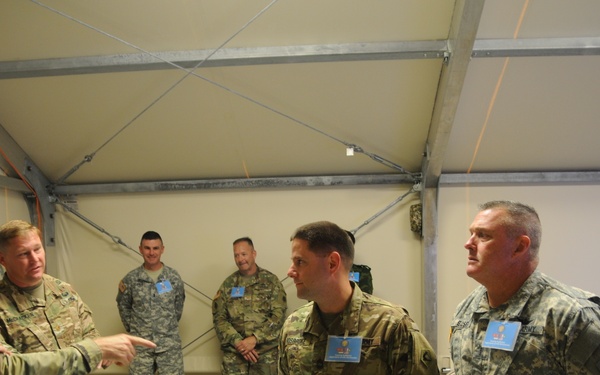 A group of approximately seven U.S. Army personnel in camouflage uniforms are gathered inside a temporary structure or tent facility. A senior officer on the left appears to be briefing or directing the group, gesturing toward others. Several personnel are wearing blue identification badges, suggesting a formal exercise, training event, or command visit.