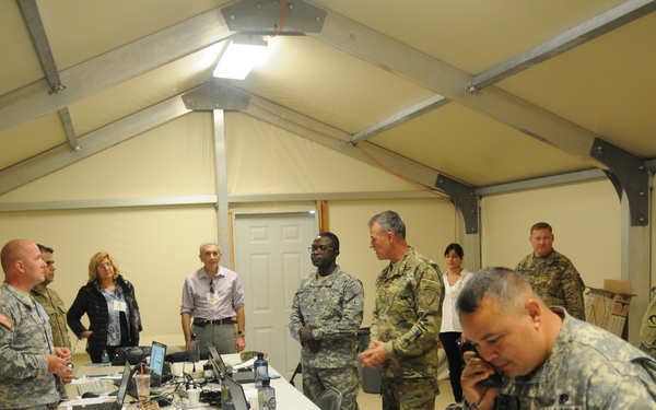 A military command post or operations center set up inside a temporary tent structure, with multiple U.S. Army personnel and at least two civilians gathered around a table covered with laptops, cables, and documents. One soldier in the foreground is speaking on a telephone while others appear to be in discussion or briefing. The setting suggests a field operations center or tactical command post during a training exercise or real-world operation.