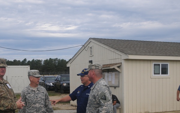 A group of military personnel are gathered outdoors at what appears to be a military installation or training facility. Four individuals in the foreground are engaged in conversation, including personnel in Army combat uniforms and one individual in a U.S. Coast Guard uniform. Two additional individuals are visible in the background near a building.
