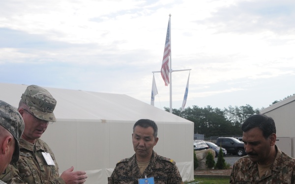 A group of military personnel from multiple nations are gathered outdoors near white tent structures, appearing to be engaged in a briefing or discussion. Visible insignia indicate the presence of US Army soldiers, a Pakistan Army officer, and what appears to be a Mongolian military officer. The setting suggests a multinational military exercise or conference.
