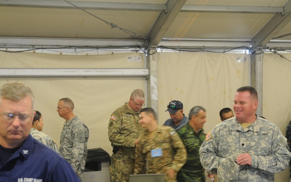 A multinational military coordination center operating inside a large tent or temporary structure. Personnel from multiple nations and branches, including U.S. Army, U.S. Coast Guard, and Mongolian military, are working at laptop-equipped tables. The setting appears to be a joint exercise or operations center with numerous uniformed and civilian participants.