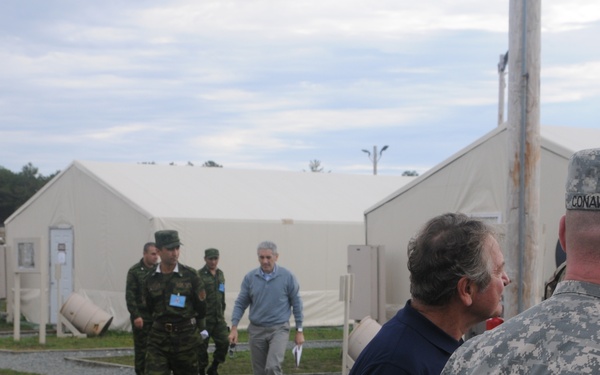 The image shows a military installation or forward operating base with multiple white tent structures. A mixed group of military personnel in different uniforms and civilians are gathered, appearing to conduct a tour or inspection of the facility. At least two distinct military uniform types are visible, suggesting a multinational or joint exercise environment.