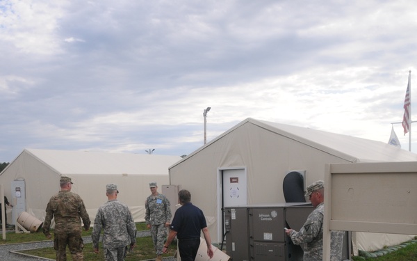The image shows a group of military personnel and at least one civilian walking through a temporary military installation consisting of large white tent structures on a gravel pathway. Several individuals are carrying cylindrical containers, suggesting setup or logistics activity. An American flag and additional flags are visible in the background near the tent structures.