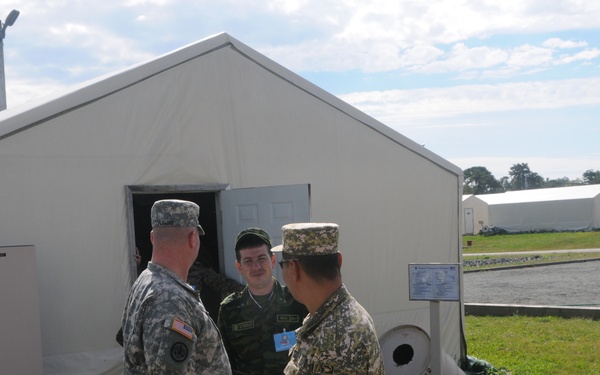 Three military personnel in different national uniforms are conversing outside a large white military tent structure at what appears to be a multinational military installation or training facility. The soldiers wear distinctly different camouflage patterns suggesting they represent at least two or three different nations. The setting includes multiple similar tent structures visible in the background, indicating a temporary military camp or exercise area.