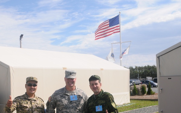 Three military personnel in different uniforms pose together outdoors at what appears to be a multinational military installation or exercise. An American flag is prominently displayed on a flagpole in the background, along with at least one additional flag. The soldiers are giving thumbs-up gestures and appear to be in a cooperative, friendly setting.