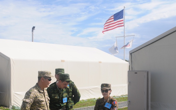 Three military personnel in different camouflage uniforms are conversing outside a temporary military installation. A US flag and two other flags are visible flying in the background, suggesting a multinational military exercise or joint operation. The setting includes modular tent structures and a numbered building (3665), consistent with a forward operating base or training camp.