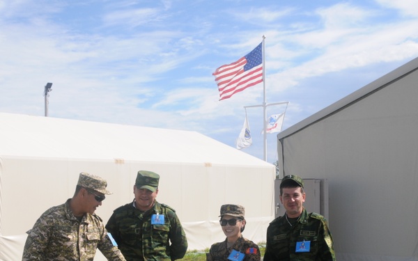 Four military personnel in different camouflage uniforms are posing together at what appears to be a multinational military installation or exercise base. An American flag and at least one other flag are visible flying in the background. The setting includes military-style tent structures and a numbered building (3665), suggesting a temporary or forward operating base environment.