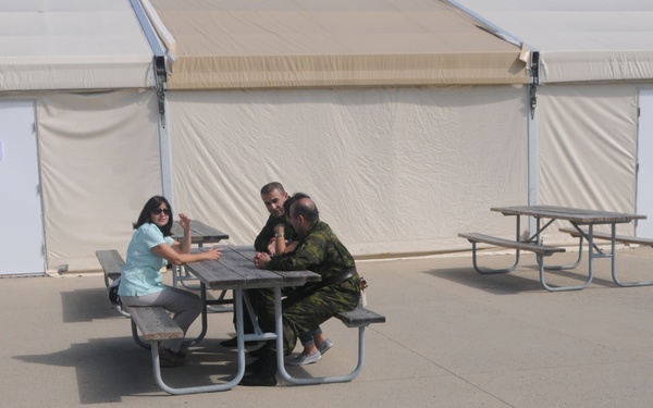 Three individuals are seated at an outdoor picnic table on a paved surface in front of a large white tent structure with multiple doors. Two of the individuals are wearing military camouflage uniforms, while the third is dressed in civilian clothing (light blue top). The setting appears to be a temporary military or emergency operations facility.