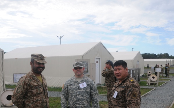 Three military personnel in different uniforms are posing together at what appears to be a military base or training facility with large white tent structures. The soldiers appear to represent different nations based on their distinct camouflage patterns and insignia. The setting suggests a multinational military exercise or joint training event.