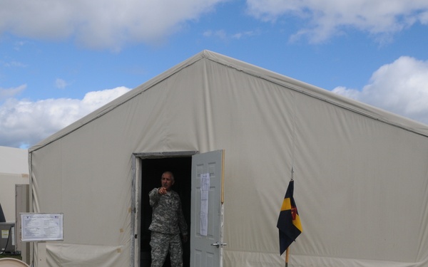 A military personnel in ACU (Army Combat Uniform) stands in the doorway of a large tan military tent structure, pointing toward the camera. The scene appears to be a forward operating base or military training installation with multiple similar tent structures visible in the background. A unit guidon flag is planted near the entrance on a gravel pathway.