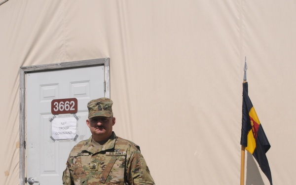 A U.S. Army soldier in OCP (Operational Camouflage Pattern) uniform stands in front of a large tan military tent structure numbered 3662, with a sign reading '51st Troop Command' on the door. A unit guidon flag on a pole is visible to the right of the soldier. The setting appears to be a military installation or field operations area.