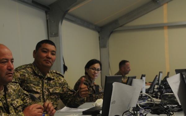 Military personnel in camouflage uniforms are seated at a long table in what appears to be a temporary command or operations center. They are working at laptops and reviewing documents, suggesting a training exercise, briefing, or joint military operation. The setting includes multiple workstations with computers, cables, and paperwork spread across the table.