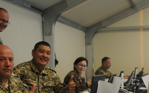Military personnel from multiple nations are seated at a long table equipped with laptops and desktop computers in what appears to be a temporary command or training facility. Two soldiers in the foreground are giving thumbs-up gestures toward the camera, suggesting a positive or successful exercise. The setting appears to be a multinational military exercise or training event.
