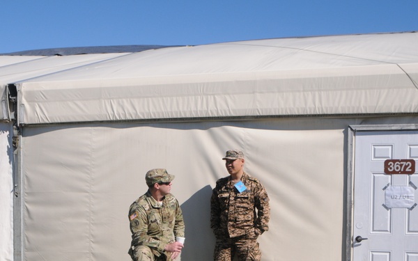 Two military personnel in different camouflage uniforms are conversing outside a large military tent structure numbered 3672. The setting appears to be a forward operating base or military installation, with sandbags lining the base of the tent structure. A third soldier in camouflage is partially visible in the foreground right.