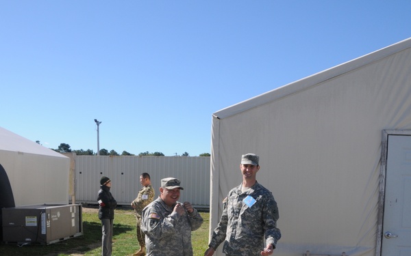 Two U.S. Army soldiers in ACU (Army Combat Uniform) camouflage stand in conversation near the entrance of a large military tent structure. The setting appears to be a forward operating base or field exercise area with multiple tent structures and equipment visible. Two additional individuals are visible in the background near what appears to be HVAC or generator equipment.