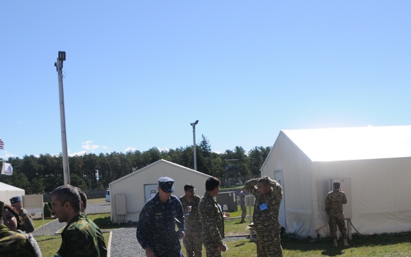 The image shows a military installation or training facility with personnel in various uniforms gathered outdoors. Multiple soldiers wearing different camouflage patterns are present, suggesting a multinational or joint military exercise. The setting includes temporary white tent structures, a gravel pathway, and an American flag visible in the background.