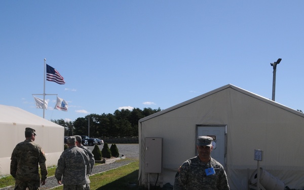 The image shows a military installation or forward operating base with several soldiers in U.S. Army camouflage uniforms moving about a gravel pathway area. The facility consists of modular tent-style structures with a U.S. flag and two additional flags flying on a flagpole in the background. The setting appears to be a training or operational base during daytime under clear blue skies.